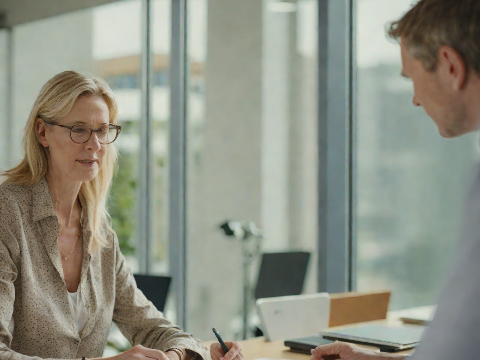 A reviewer slides a single printed page across a table to a business owner, with a software team at monitors visible behind a glass wall.